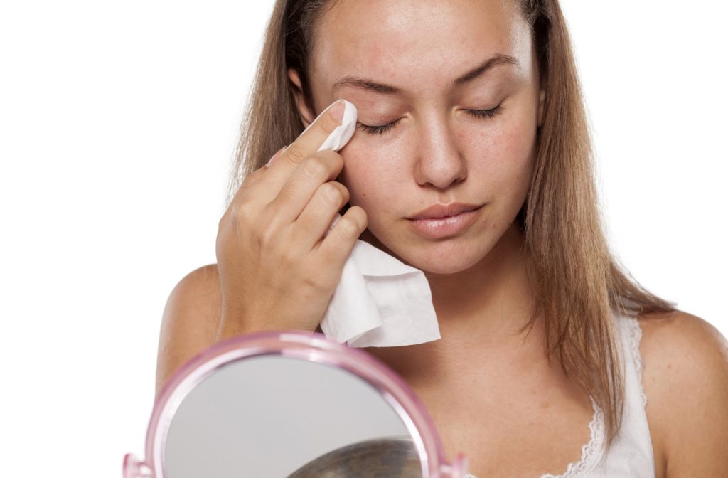 Young woman using face cloth to wipe around eyes while using mirror.