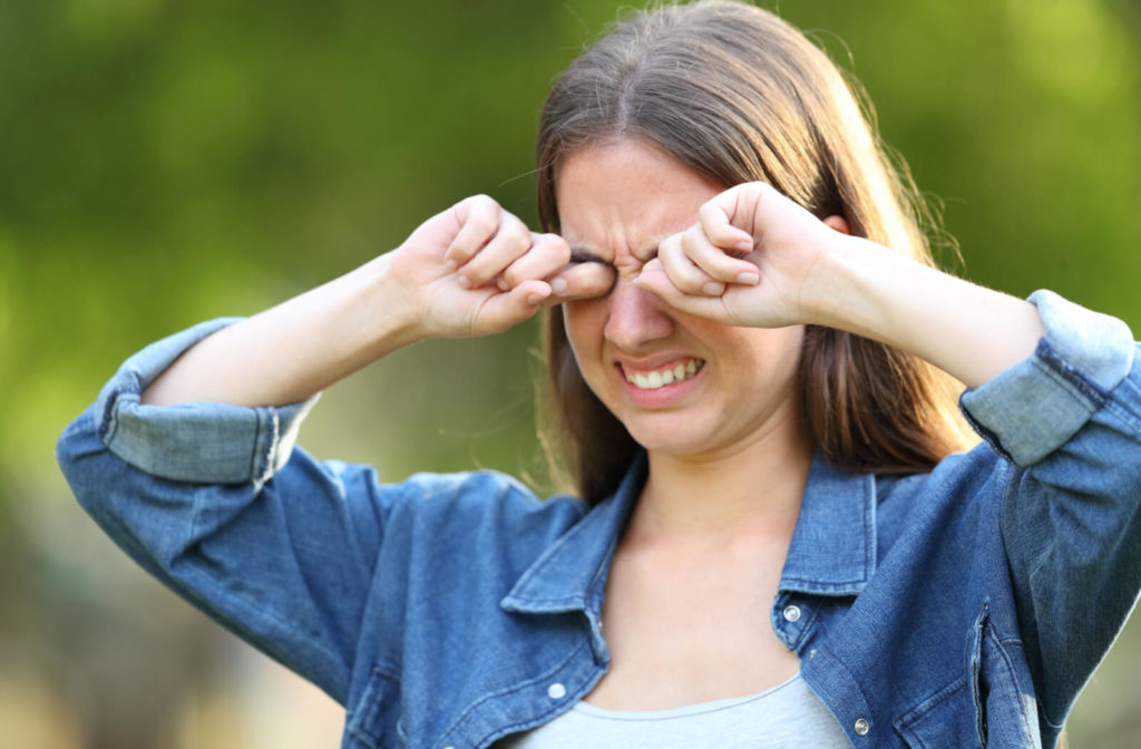 A girl in a denim shirt is rubbing her eyes with both hands and showing irritation on her face.