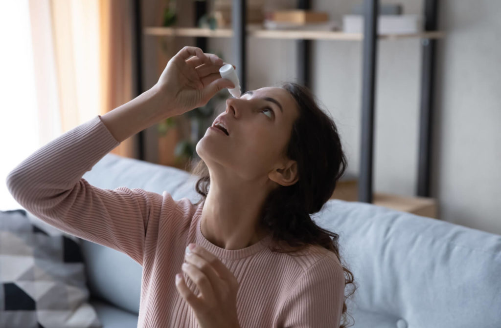 A lady sitting on a sofa is holding a bottle of prescribed eye drops close to her right high and pouring some drops.