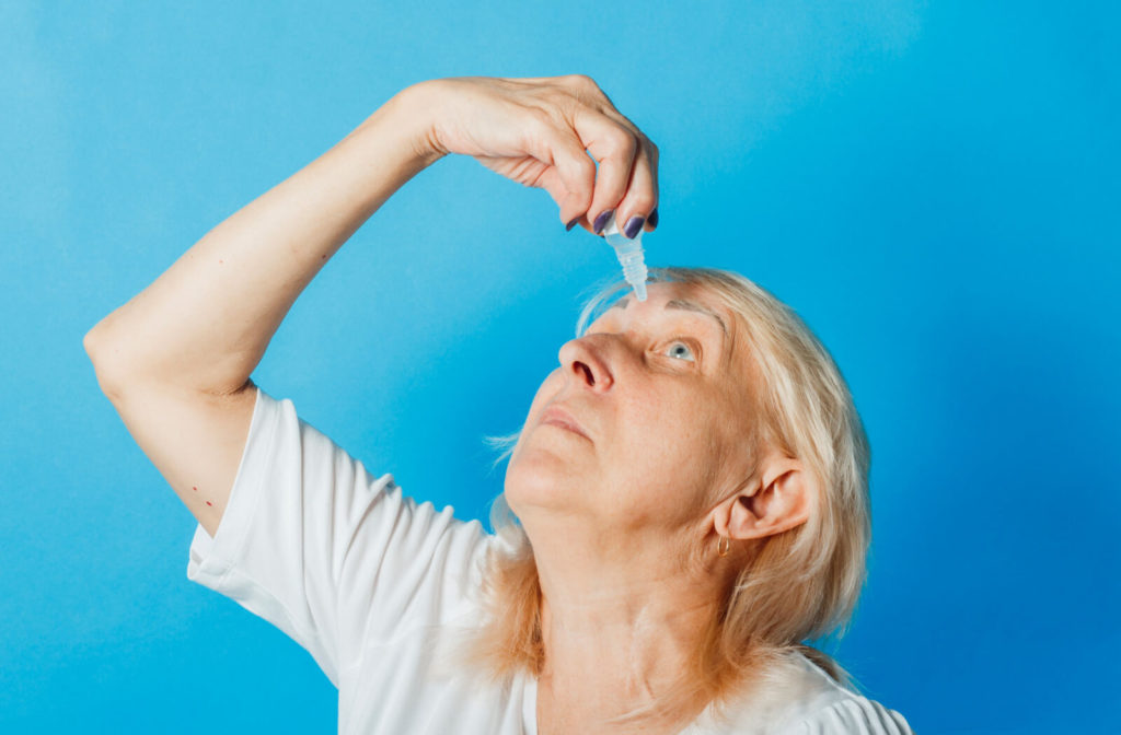 a woman against a blue background applies eye drops to her left eye 