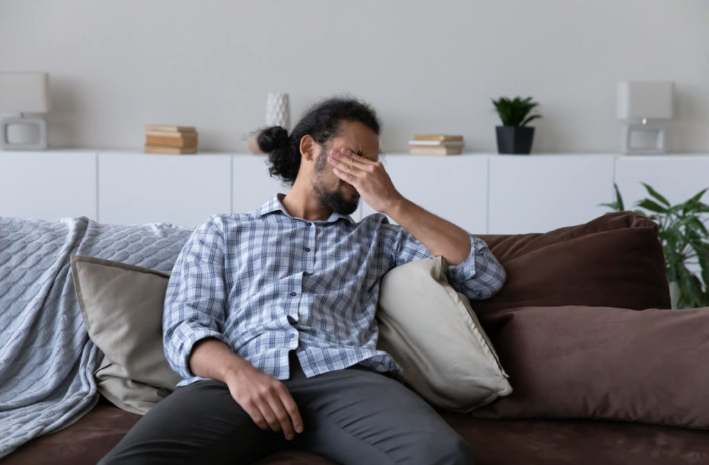 A man sitting on a couch and holding his left hand over his eyes, squinting as if in pain