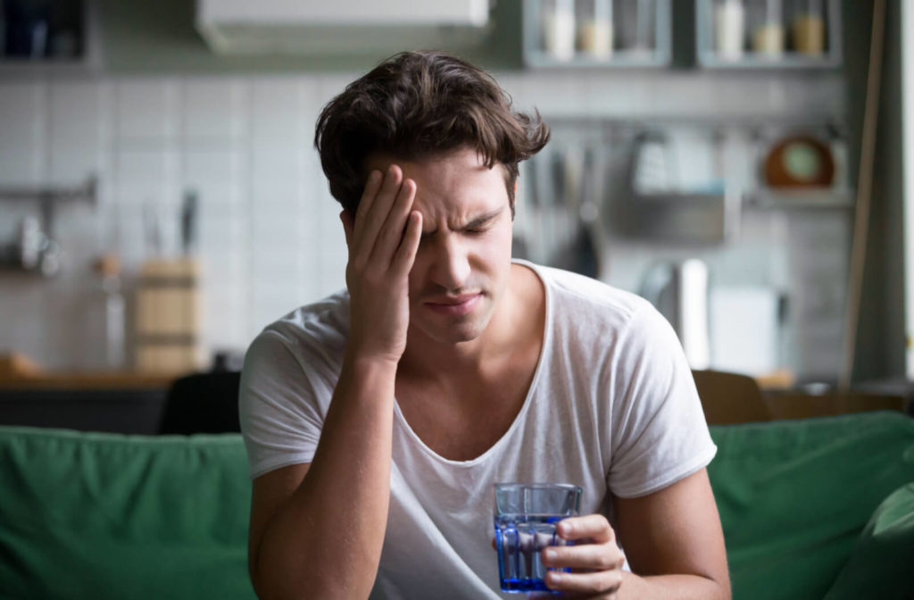 Close-up of a young man sitting on a couch and holding a glass of water as he closes his eyes and presses his right hand to his forehead