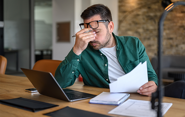 White man is looking stressed, and they are rubbing their eyes and bridge of their nose. They are sitting at a desk full of paperwork and a laptop.