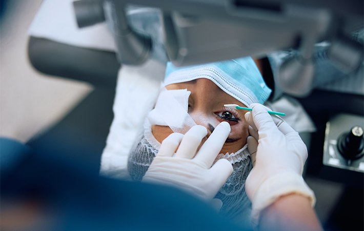 Women undergoing laser eye surgery with one eye opened up wide. Other eye is covered by guaze. A doctor's hands are seen cleaning the eye of the operation.