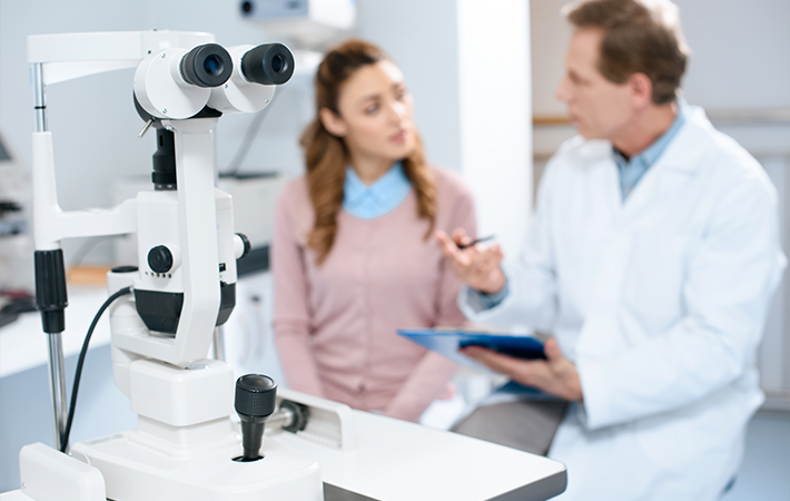 A Slit Lamp is in the foreground of an optometrist office. In the background there is a male doctor talking to a woman patient.