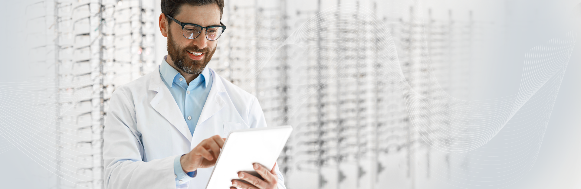 Male optometrist doctor, holding a white tablet, smiling and scrolling. There is a wall of glasses in blurred in the background.
