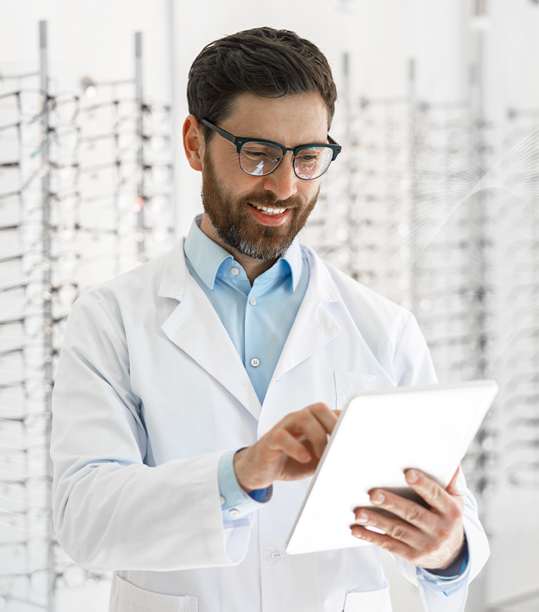 Male optometrist doctor, holding a white tablet, smiling and scrolling. There is a wall of glasses in blurred in the background.